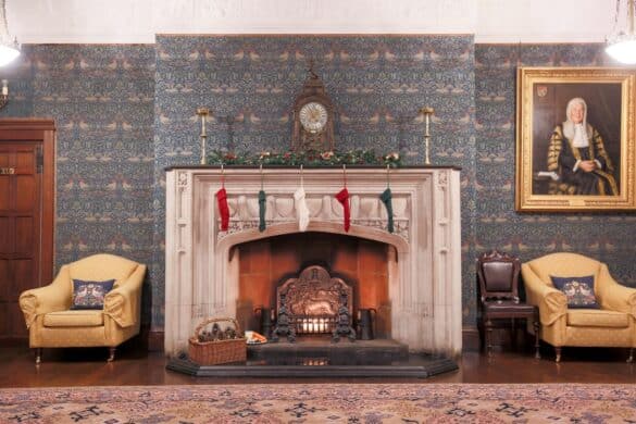 A fireplace in a Victorian wallpapered room at Ironmongers Hall, London, with red, white and green Christmas stocking hanging from the mantelpiece that holds Christmas decorations and an antique brass clock