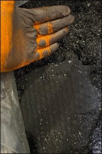 A hand in a yellow glove pointing at a pile of black mass, a battery residue containing valuable metal, that has a mixture of soil-like compacted and loose textures