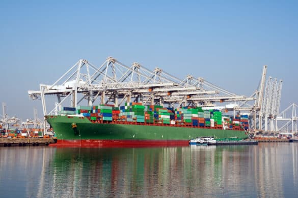 Image of a cargo ship at port loaded with containers, with the wharf and port cranes in eh background