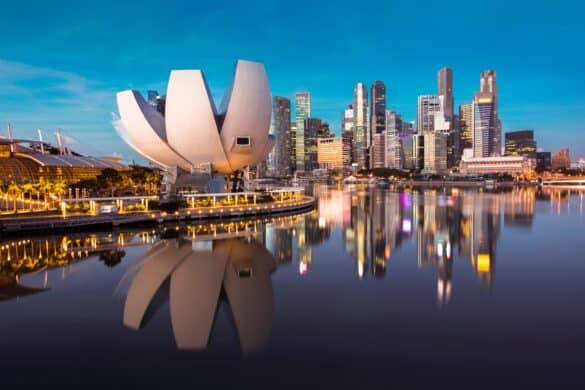 Waterside view of Singapore at dusk, featuring cityscape, its reflection in the water and a bright blue sky. Photo by MOLPIX at Shuttlestock