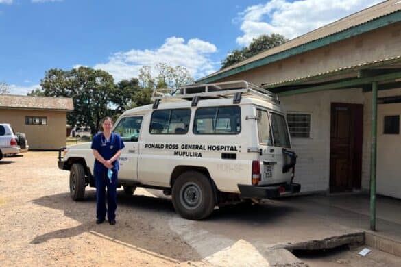 Photo of a girl ith mid-brown haor, wearing a medical uniform sanding next to an ambulance with text on its side saying 'Ronald RossGeneral Hospital' at a building entrance