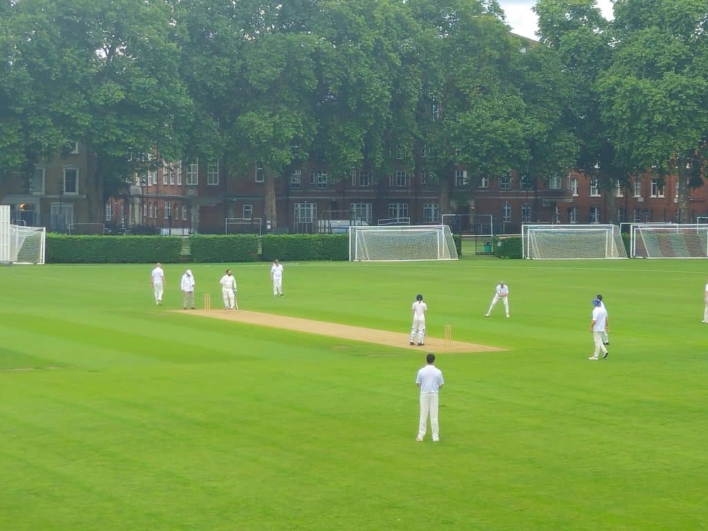 A game of cricket on a grass playing field, with trees and a London terrace of Victorian houses in the background