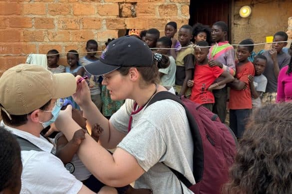 Medical students in white t-shirts and baseball caps examining a child patient, with other children in the background