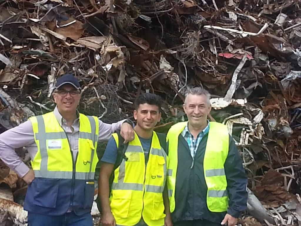 Three men in high visibility jackets standing together with a scrap metal pile behind them