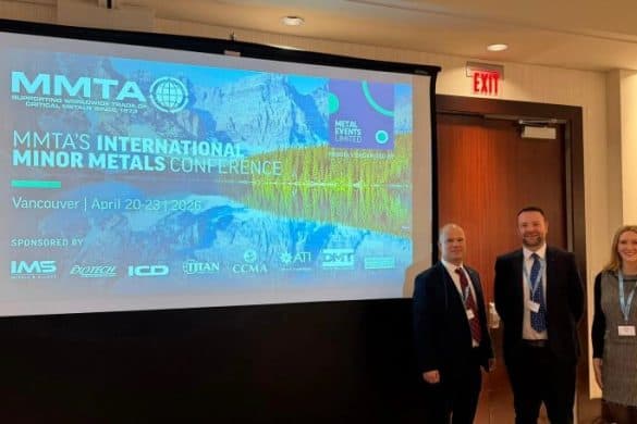 Standing by the MMTA International Minor Metals Conference 2026 display screen in the conference room at Hyatt Regency Vancouver, left to right: MMTA outgoing chairman Stephen Hall, a close-shaven man in a dark suit, newly-elected chairman Chris Brzozowski, a man with dark hair and a trim beard, wearing a dark suit, and Freya Kerwin,MMTA managing director, a young woman with long blond hair wearing a grey pinafore dress and a black top.