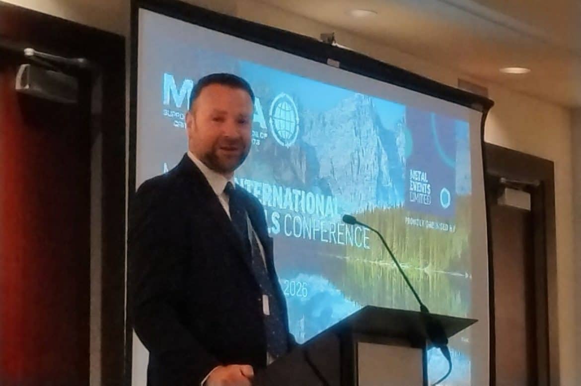 MMTA chairman Chris Brzozowski, a dark haired man with a trim beard speaking at a podium in front of an MMTA conference screen.