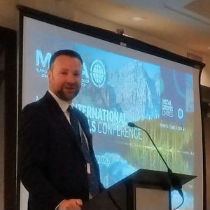 MMTA chairman Chris Brzozowski, a dark haired man with a trim beard speaking at a podium in front of an MMTA conference screen.