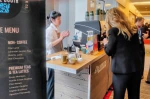 A coffee station with a display next to it, a barista and a customer, with others in the background.