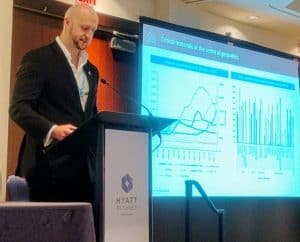 Scott Yarham, head of strategy at Project Blue, a tall man with a short beard is speaking at a podium, with charts on a conference screen in the background. 