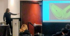 Theo Ruas of Indium Corp, a man with long dark hair and glasses, wearing a dark jumper is speaking in a conference troom, pointing at a screen with an illustration of the butterfly effect in green on a black background.